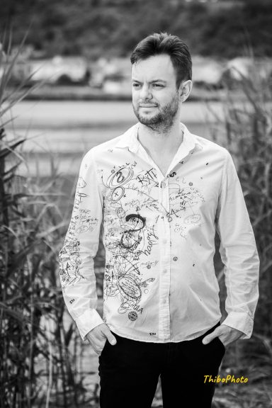 Homme posant en chemise blanche avec motifs, entouré de végétation.