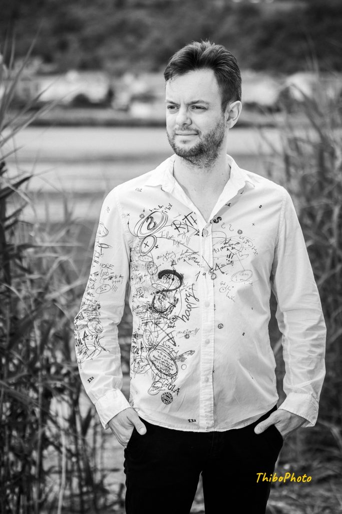 Homme posant en chemise blanche avec motifs, entouré de végétation.