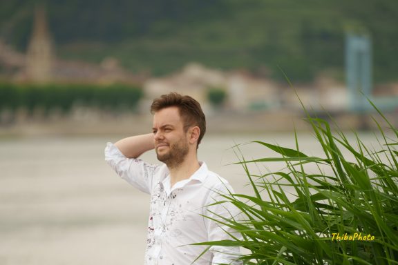 Homme souriant en chemise blanche près d'une rivière, entouré de verdure.