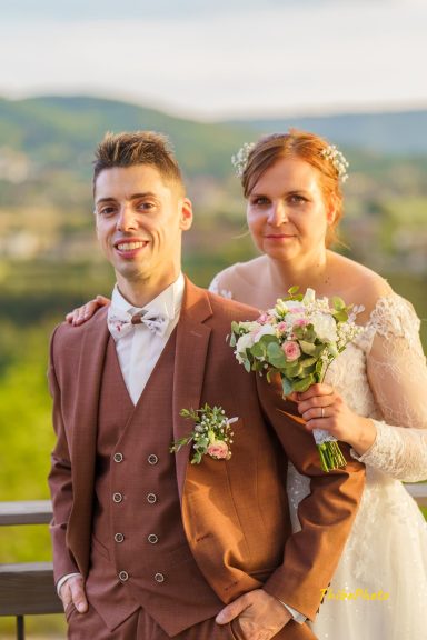 Jeune couple heureux en tenue de mariage, avec des fleurs, devant un paysage verdoyant.