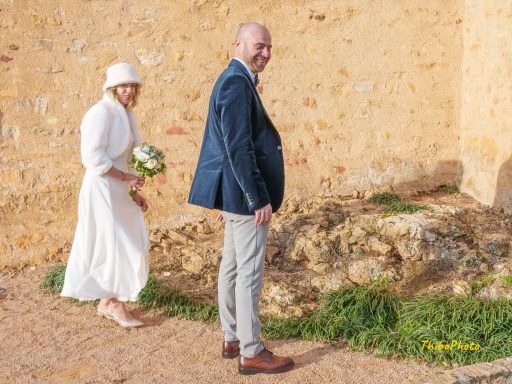 Un couple souriant marche sur un chemin en pierre, devant un mur ensoleillé.