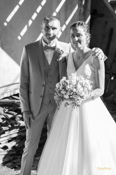 Un couple de mariés sourit, debout ensemble, fleurs à la main, dans un cadre romantique.