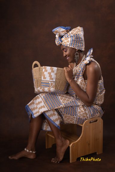 Femme assise, souriante, vêtue d'un pagne, tenant un sac en tissant des fils.