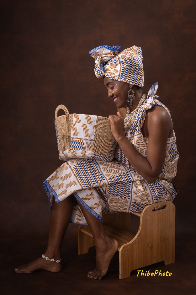 Femme assise, souriante, vêtue d'un pagne, tenant un sac en tissant des fils.