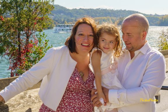 Une femme, un homme et une petite fille sourient au bord de l'eau, entourés de verdure.