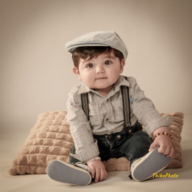 Un jeune enfant assis sur un coussin, vêtu d'une chemise et d'un chapeau gris.