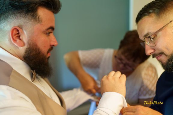 Deux hommes en costume s'habillent, tandis qu'une femme ajuste un détail.