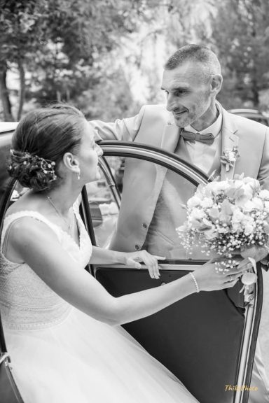 Un couple de mariés souriant devant une voiture, la mariée tenant un bouquet.