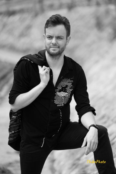 Homme souriant, habillé en noir, posant sur le sable avec un bras sur la cuisse.