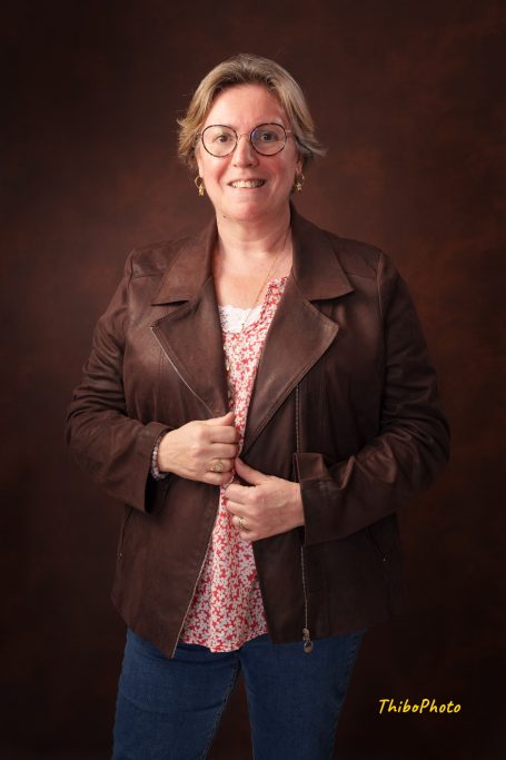 Femme souriante en veste en cuir marron, blouse rose sous la veste, fond marron.
