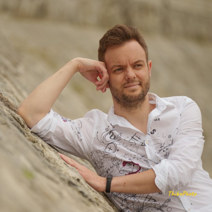 Homme souriant en chemise blanche, reposant sur une surface rocheuse.