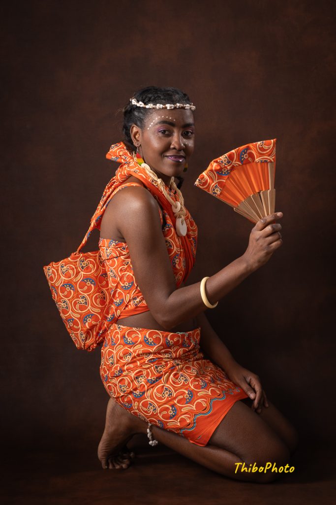 Femme en tenue traditionnelle orange, tenant un éventail, posant avec un sourire.