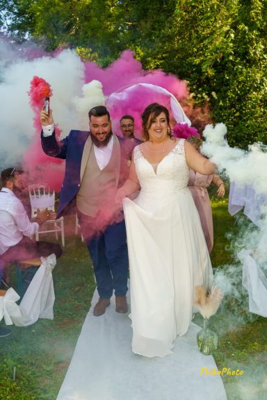 Un couple de mariés marche joyeusement avec des fumigènes colorés en arrière-plan.