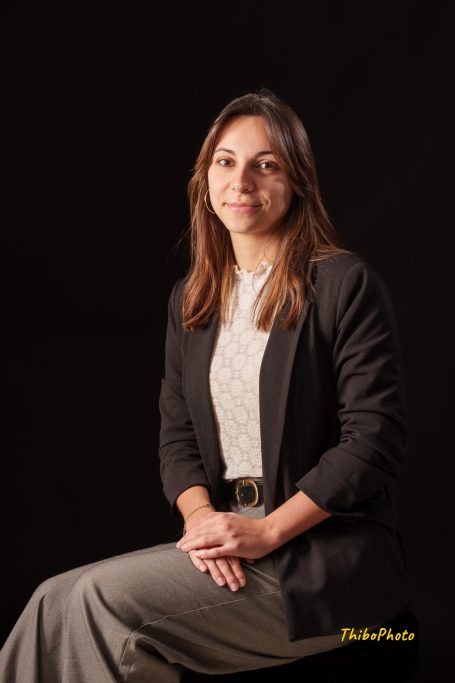 Femme souriante assise en tenue professionnelle sur fond sombre.