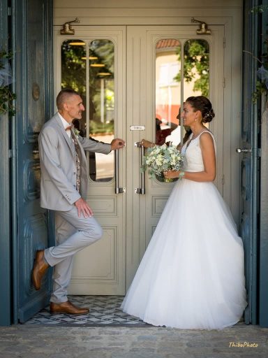 Un couple à la porte, elle en robe de mariée, lui en costume, sourire à la main.