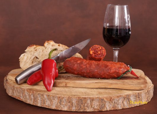 Planche en bois avec du saucisson, piments, pain et un verre de vin rouge.