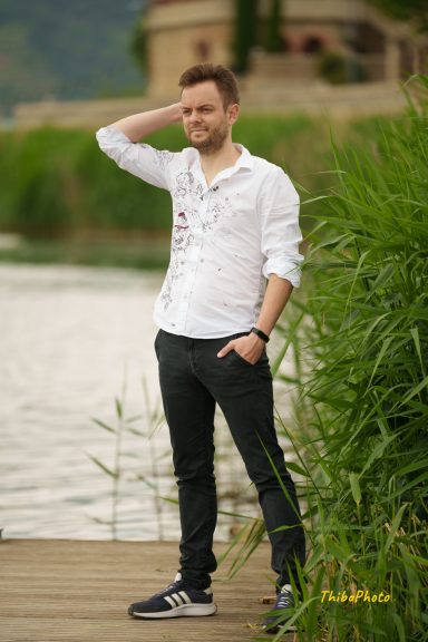 Homme souriant en chemise blanche et pantalon noir près de l'eau, entouré de végétation.
