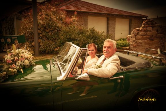 Mariage avec couple souriant dans une voiture décapotable, fleurs en arrière-plan.