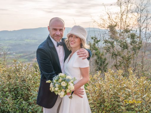 Un couple souriant, habillé élégamment, posant avec un bouquet de fleurs en plein air.