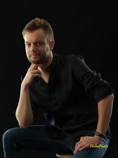 Homme pensif avec une chemise noire, assis en lumière douce sur fond noir.