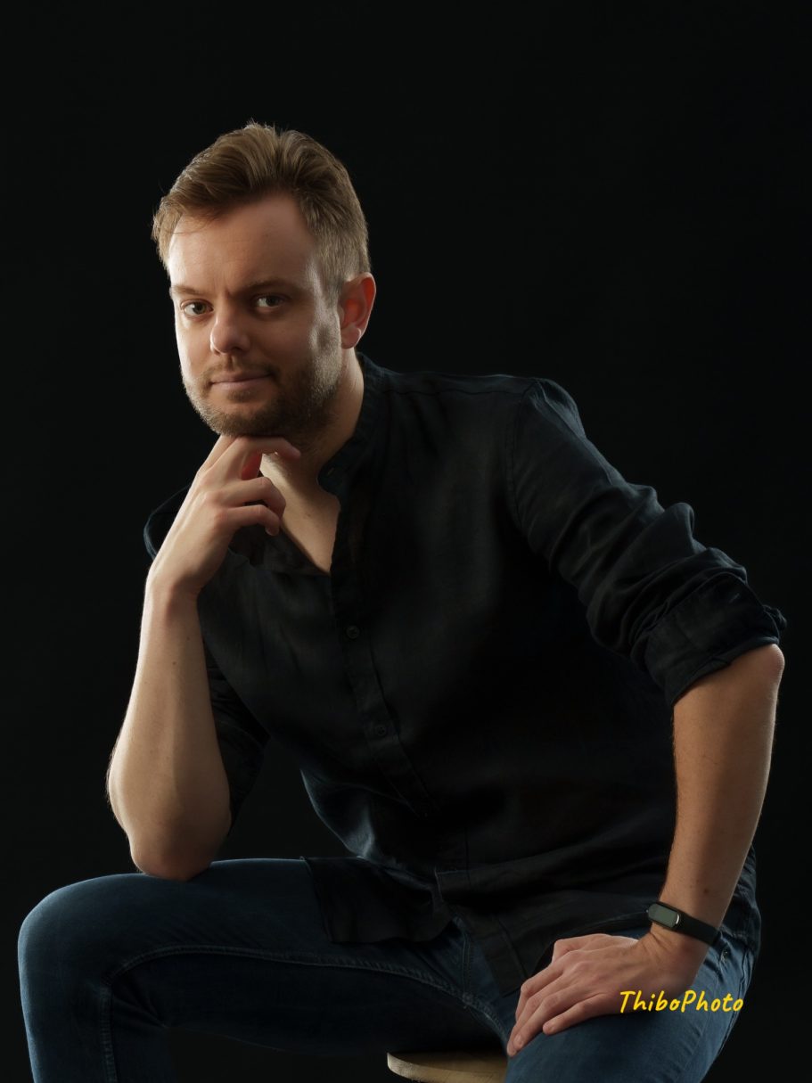 Homme pensif avec une chemise noire, assis en lumière douce sur fond noir.