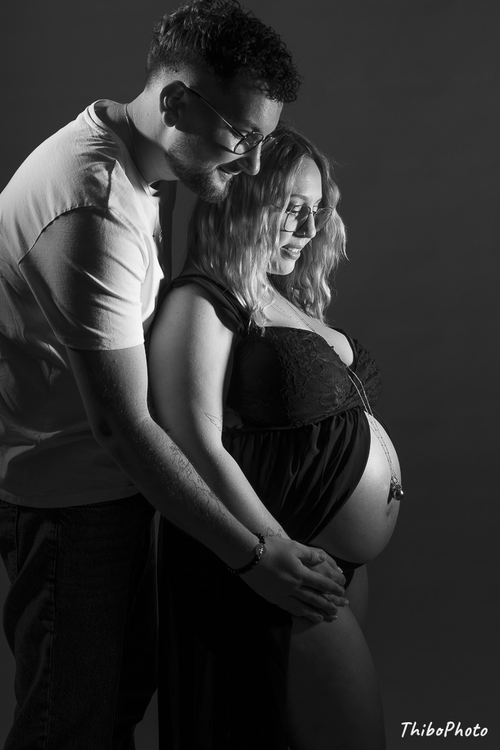 Un couple pose en noir et blanc, mains sur un ventre de femme enceinte.