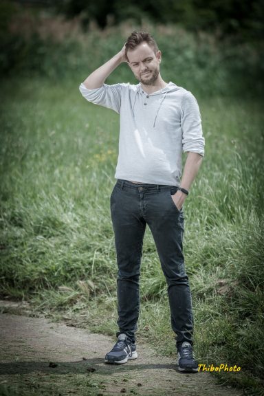 Homme debout dans un champ, portant un pull gris et un pantalon foncé, sourire léger.