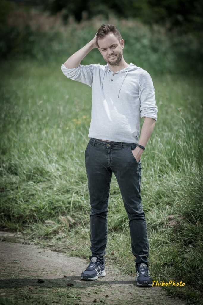 Homme debout dans un champ, portant un pull gris et un pantalon foncé, sourire léger.