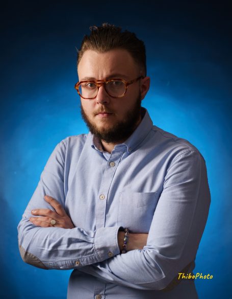 Homme avec lunettes, chemise bleu clair, bras croisés, fond bleu.