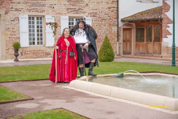 Deux personnes en costumes médiévaux se tiennent près d'une fontaine dans un jardin.